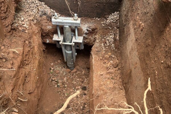 Galvanized helical pile bracket under a brick foundation footing in a hand-dug hole with red clay and roots