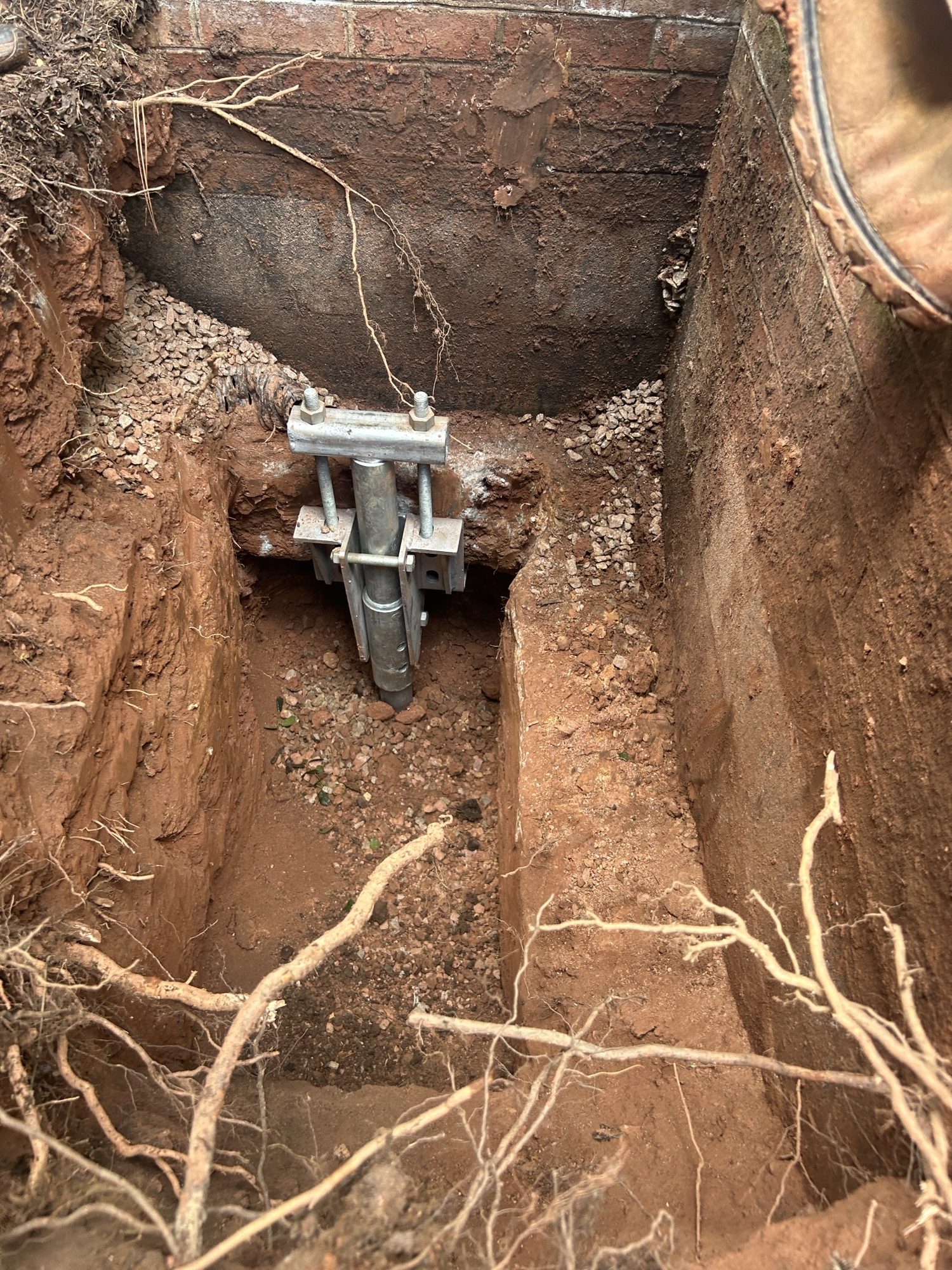 Galvanized helical pile bracket under a brick foundation footing in a hand-dug hole with red clay and roots